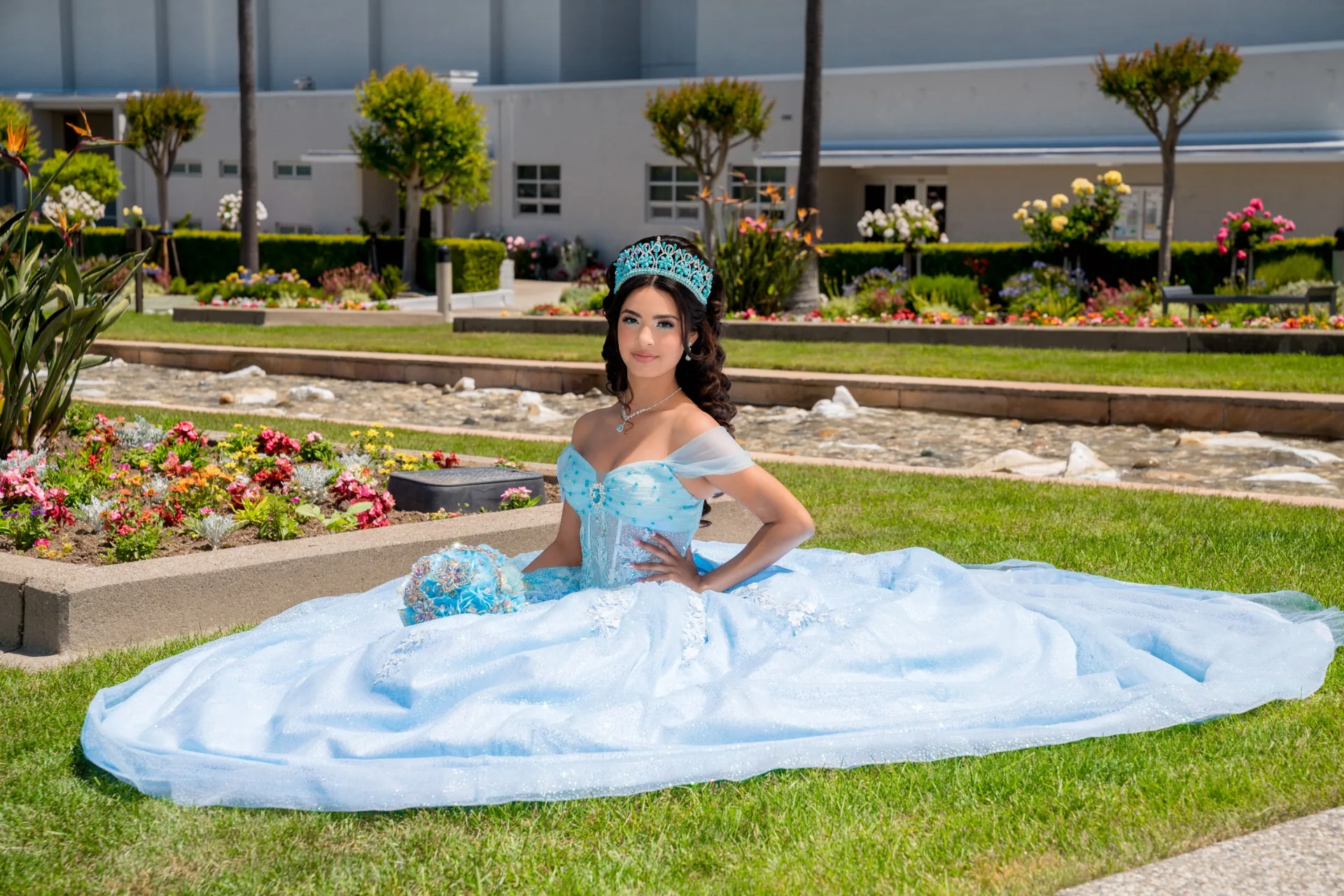 Quinceañera sentada con el vestido celeste extendido sobre el césped de los jardines